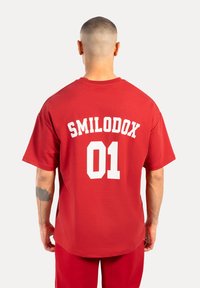 Camiseta de algodón roja con un gran estampado blanco en la parte posterior que dice "SMILODOX 01". Mangas cortas y un corte holgado.