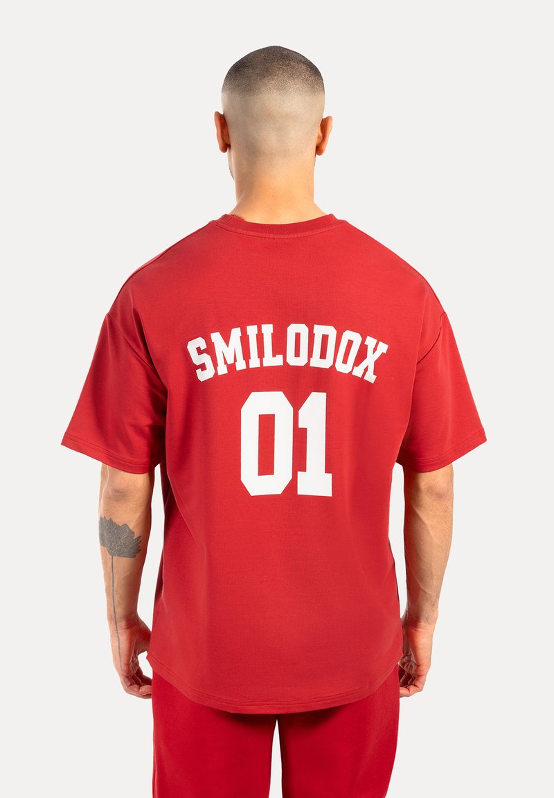 Camiseta de algodón roja con un gran estampado blanco en la parte posterior que dice "SMILODOX 01". Mangas cortas y un corte holgado.