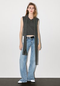 Grijze mouwloze gebreide top met een V-hals, gecombineerd met lichtblauwe flared jeans en een lange grijze sjaal die naar beneden hangt. Witte schoenen maken de look compleet.