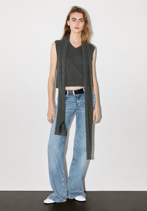 Top en tricot sans manches gris avec un décolleté en V, associé à un jean évasé bleu clair et à une longue écharpe grise tombante. Des chaussures blanches complètent le look.