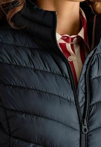 Ciemnoniebieska puchowa kurtka z wysokim kołnierzem, z poziomym pikowaniem, z zapięciem na zamek z przodu i kontrastową koszulą w paski widoczną pod spodem.