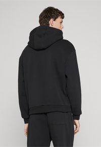 Uomo di spalle che indossa una felpa con cappuccio nera semplice e pantaloni neri abbinati su uno sfondo grigio chiaro.