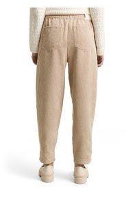 Personne portant un pantalon beige en velours côtelé avec une taille élastique et des poches arrière, associé à un pull en maille crème et des chaussures beiges, debout de dos.