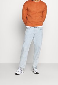 Pomarańczowy dziany sweter, jasnoniebieskie luźne jeansy oraz białe trampki z fioletowymi akcentami. Prosty design z teksturowaną tkaniną swetra.