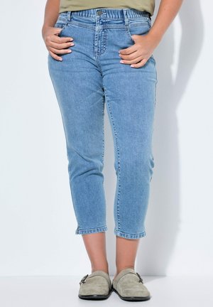 Kvinde iført lyseblå korte jeans med lommer, beige slip-on sko og en lysegrøn top, delvist synlig. Hænderne i lommerne.