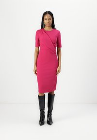 Robe ajustée magenta avec des manches courtes et un design drapé, associée à des bottes noires jusqu'aux genoux, sur un fond blanc uni.