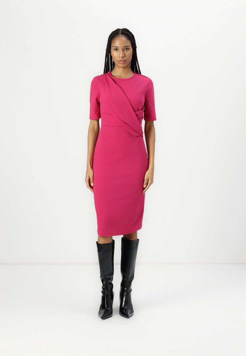 Robe ajustée magenta avec des manches courtes et un design drapé, associée à des bottes noires jusqu'aux genoux, sur un fond blanc uni.
