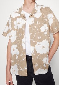 Camicia a maniche corte in stampa floreale beige e bianca, realizzata in tessuto traspirante. Presenta un colletto e una tasca frontale, con una vestibilità comoda.