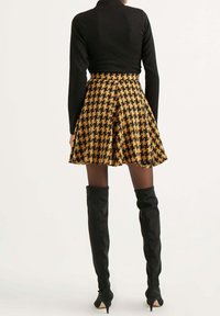 Mujer vestida con una blusa negra de manga larga, falda de línea acampanada de cuadro de galés en marrón y negro, medias translucidas y botas altas de tacón negras, de espaldas.