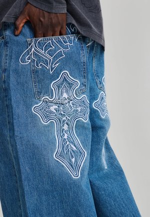 Hand ruht in der Gesäßtasche einer blauen Jeans mit weißem, besticktem Kreuz und floralen Designs am Bein vor einfarbigem Hintergrund.