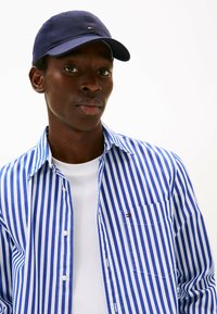 Camisa de botones a rayas azul y blanca con un bolsillo en el pecho; combinada con una camiseta blanca lisa y una gorra navy con un pequeño logo.