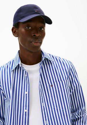 Chemise à rayures bleues et blanches à boutons avec une poche poitrine ; associée à un T-shirt blanc uni et une casquette marine avec un petit logo.