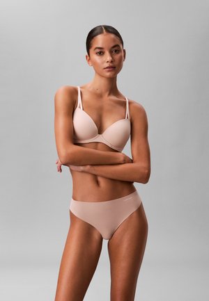 Femme debout, bras croisés, portant un soutien-gorge beige clair et une lingerie assortie sur fond gris uni.
