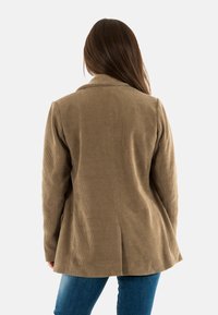 Mujer con cabello largo castaño, vistiendo una chaqueta de pana de color marrón claro y jeans azules, de espaldas contra un fondo blanco liso.