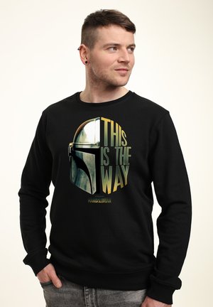 Man with short hair and ear piercings wearing a black sweatshirt featuring a Mandalorian helmet and the text "This is the way."