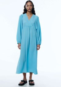 Hellblaues langes Kleid mit Wickeldesign, langen Puffärmel, strukturiertem Stoff und fließender Silhouette. Kombiniert mit schwarzen flachen Sandalen.