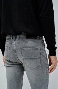 Jeans ajustados de mezclilla gris con una textura desgastada, que cuentan con bolsillos traseros y detalles de parches, combinados con una camiseta negra de manga larga.