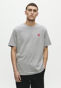 Grå bomuld t-shirt med korte ærmer, rund halsudskæring og rød logo-patch på venstre bryst. Glat tekstur og afslappet pasform.
