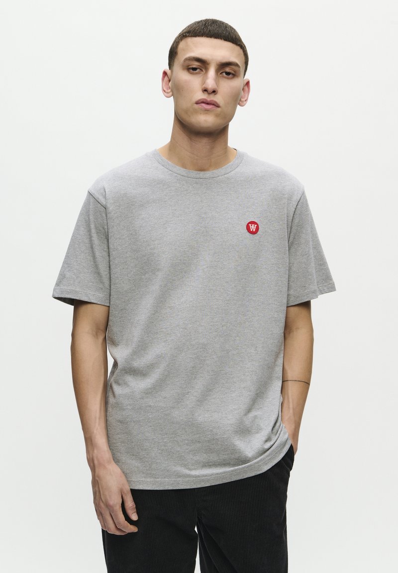 Grå bomuld t-shirt med korte ærmer, rund halsudskæring og rød logo-patch på venstre bryst. Glat tekstur og afslappet pasform.