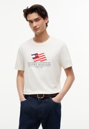Jeune homme portant un T-shirt blanc Tommy Hilfiger avec un logo drapeau rouge et marine, jean bleu, mains dans les poches, fond neutre.