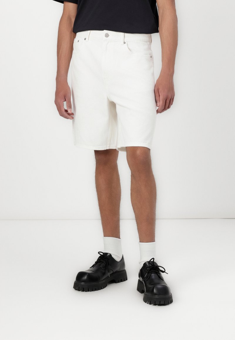 Hvide denimshorts med en klassisk pasform, der har bæltestropper og et fem-lomme-design, parret med sorte kraftige snørebåndssko og hvide sokker.