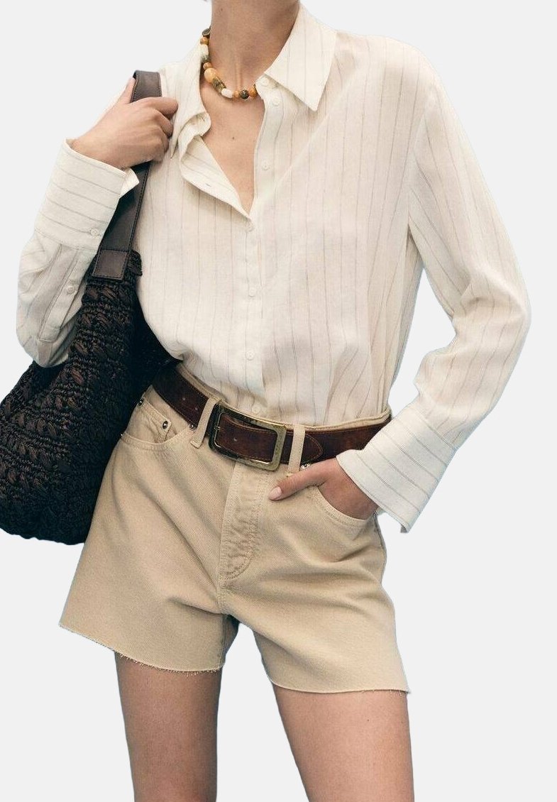 Femme portant une chemise crème rayée à boutons, un short beige avec une ceinture marron, tenant un sac sombre tissé sur une épaule.