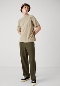 Beige katoenen t-shirt met een borstzak, gecombineerd met lichte olijfgroene broek. Zwarte sneakers met crèmekleurige rubberen zolen maken de look compleet.