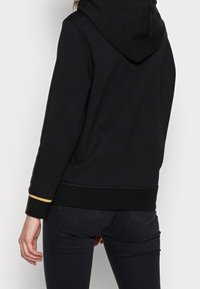 Sudadera negra con capucha que incluye un bolsillo delantero, dobladillo y puños acanalados con una franja dorada de acento. Tejido suave con un corte relajado y capucha ajustable.