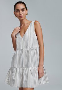 Biała sukienka z granatowymi paskami, z dekoltem w kształcie litery V, bez rękawów, z falbanami na spódnicy, wykonana z lekkiego materiału. Modelka prezentuje fason i krój.