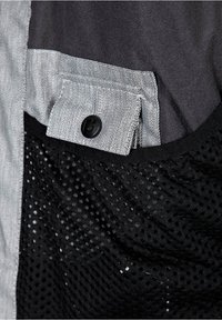 Svart nätväv ficka med en grå herringbone-flik och knappstängning, med lager av texturer och kontrast mellan materialen.