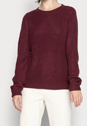 Osoba ubrana w bordowy, prążkowany sweter z dzianiny z długimi rękawami i białe spodnie, stojąca na tle jasnego, jednolitego tła.