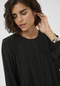Zwarte, geplooide blouse met een ronde hals en lange mouwen. Zachte textuur met een subtiele glans. Beschikt over een manchetdetail op de mouwen.