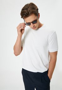 Witte gestructureerde t-shirt met korte mouwen en een ronde hals, gecombineerd met donkere broek. Persoon past zwarte zonnebril terwijl hij naar beneden kijkt.