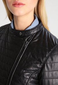 Detailní záběr na ženu, která má na sobě černou prošívanou koženou bundu se zipem a límcem na patentky, pod ní modrou košili.