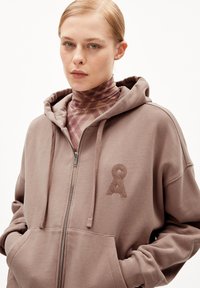 Haut de sweat à capuche zippé marron avec une texture douce, présentant une poche avant et un logo brodé. Porté par-dessus un haut à motifs avec col montant.