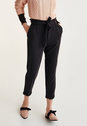 Femme portant un pantalon noir taille haute fuselé avec ceinture nouée en nœud, chevilles retroussées, pull rose en maille torsadée, et ballerines beige avec des nœuds noirs.