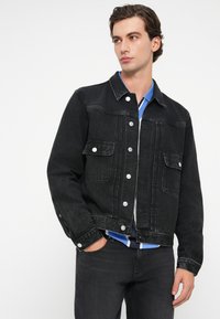 Μαύρο τζάκετ από τζιν με κλασική γραμμή, διαθέτοντας ασημένια κουμπιά κλεισίματος, δύο μπροστινές τσέπες και αντίθετες ραφές.