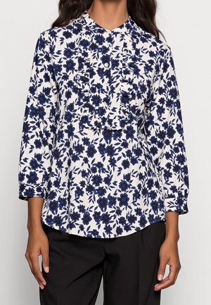 Blouse florale blanche avec des fleurs bleu marine, présentant un devant volé, des manches longues et un col rond. Portée avec un pantalon noir.