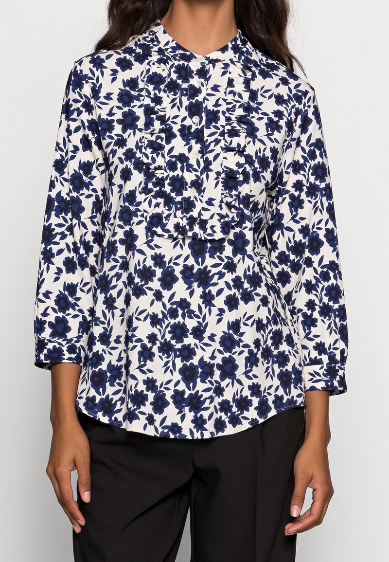 Blouse florale blanche avec des fleurs bleu marine, présentant un devant volé, des manches longues et un col rond. Portée avec un pantalon noir.