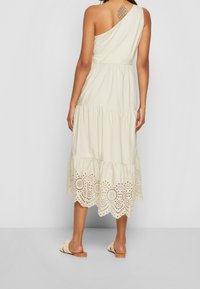 Vestido en tono beige claro con un solo hombro, con volantes en capas y un intrincado detalle recortado en el dobladillo. Diseño sencillo con un ajuste relajado.