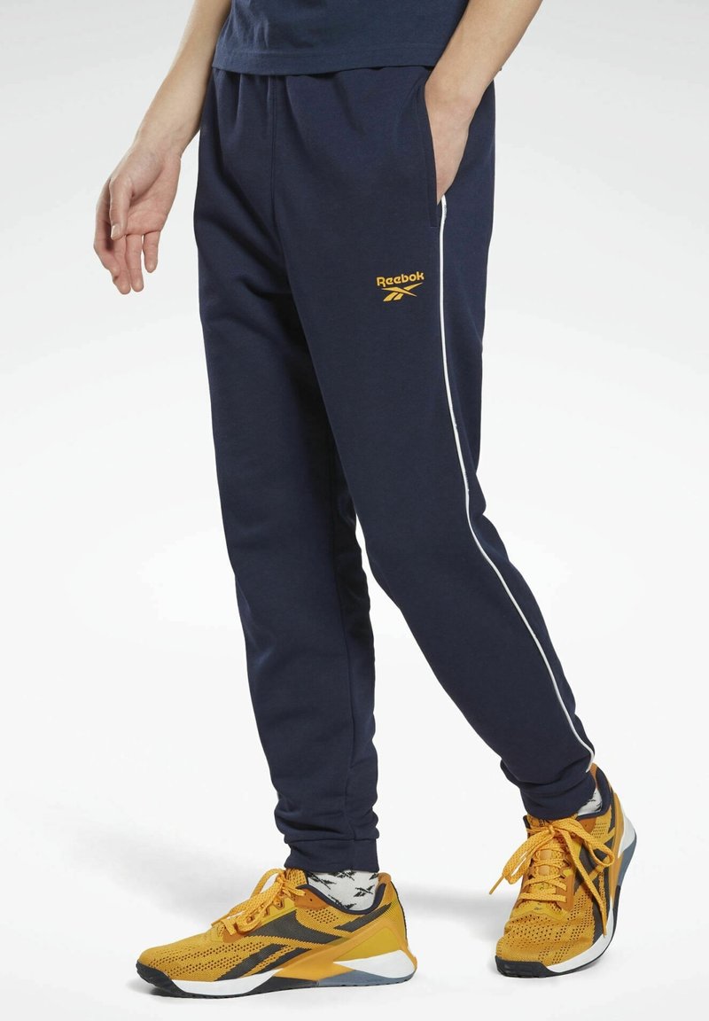 Marineblå sweatpants med hvide sidelister, Reebok-logo i gult. Fremstillet af blødt materiale, med lommer og en afslappet pasform.