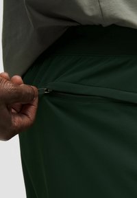 Tamsiai žali sportiniai šortai su tekstūruotu audiniu, elastinga juosta ir užtrauktuku šone, kurioje matoma ranka, traukianti užtrauktuką.