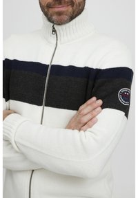 Suéter de manga larga con cremallera en blanco, que presenta rayas horizontales en navy y gris oscuro. Tejido texturizado con cuello y puños de canalé, parche con logo.