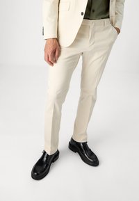 Beige dressbukser med slim fit, kombinert med svarte lærskjorter. Bukser har en glatt tekstur og minimalistisk design.