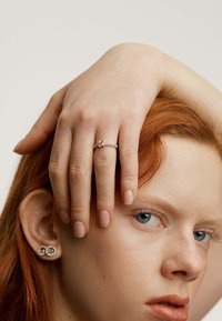 Anillo de plata con una piedra de corte esmeralda, acentuado por piedras más pequeñas y transparentes en la banda. La modelo lleva pendientes de piedras preciosas redondas.