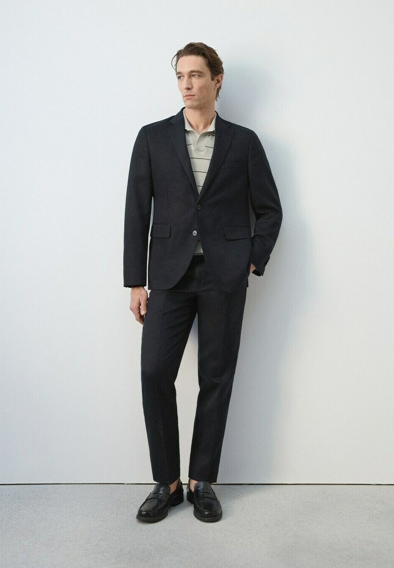 Un homme se tient contre un mur blanc, vêtu d'un costume noir, d'un polo à rayures et de mocassins noirs, une main dans la poche, regardant sur le côté.