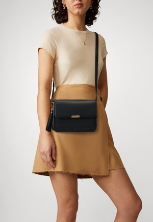 Sac bandoulière en cuir noir avec rabat, petit détail en métal doré et pompon, associé à un haut beige côtelé et une jupe marron.