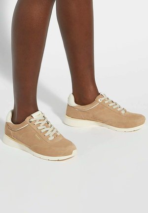 Beige suede sneakers with white accents, round toe, textured lace, and a cushioned sole. Features branding on the outer side.