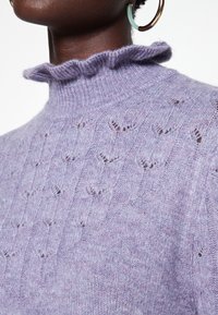 Lavendelfärgad stickad tröja med rynkad hög krage, prydd med ett fint mönster av små utskärningar. Mjuk textur, avslappnad design.