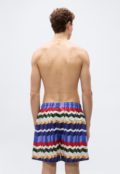 Άνδρας με σγουρά μαλλιά, όρθιος, ξυπόλητος, γυμνόστηθος, φορώντας πολύχρωμα σορτς με γεωμετρικό σχέδιο, πάνω σε απλό φωτεινό φόντο.